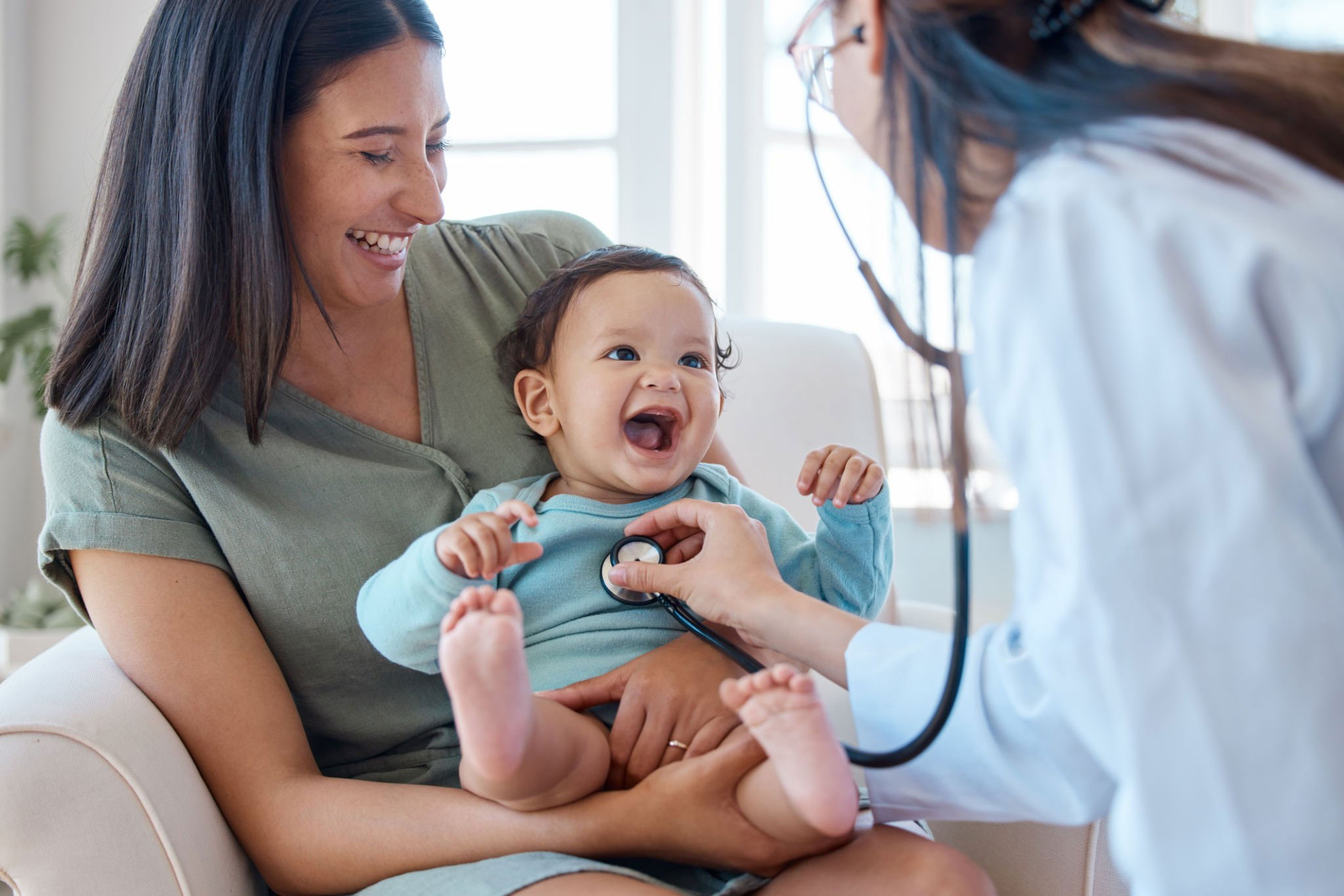 A laughing baby being examined by a smiling doctor while their mother holds them